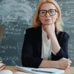 Teacher sitting at a desk with chalkboard formulas