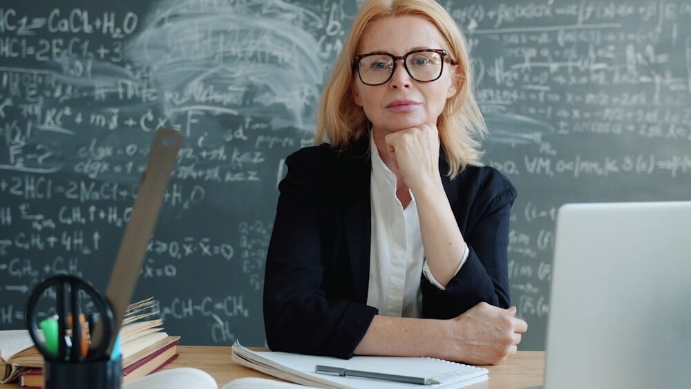 Teacher sitting at a desk with chalkboard formulas