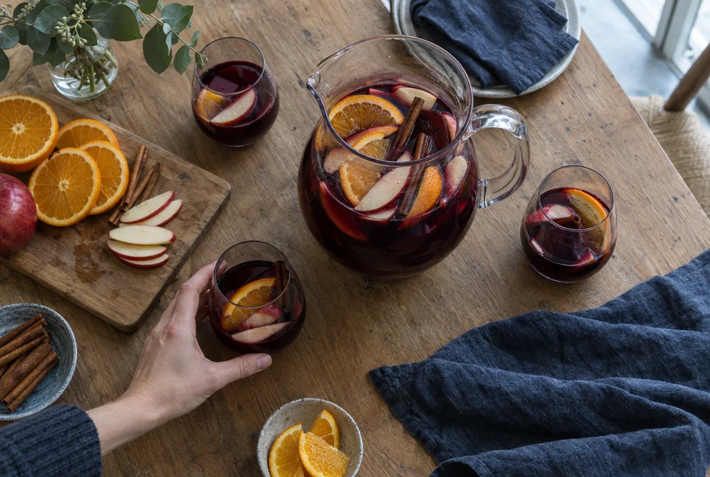 Pitcher of red sangria with orange slices, apple, and cinnamon sticks on a wooden table beside filled stemless glasses